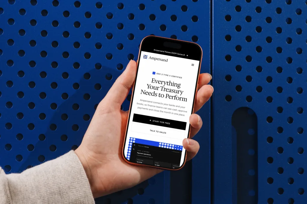 The Ampersand homepage on an iPhone held in one hand against a blue perforated cabinet