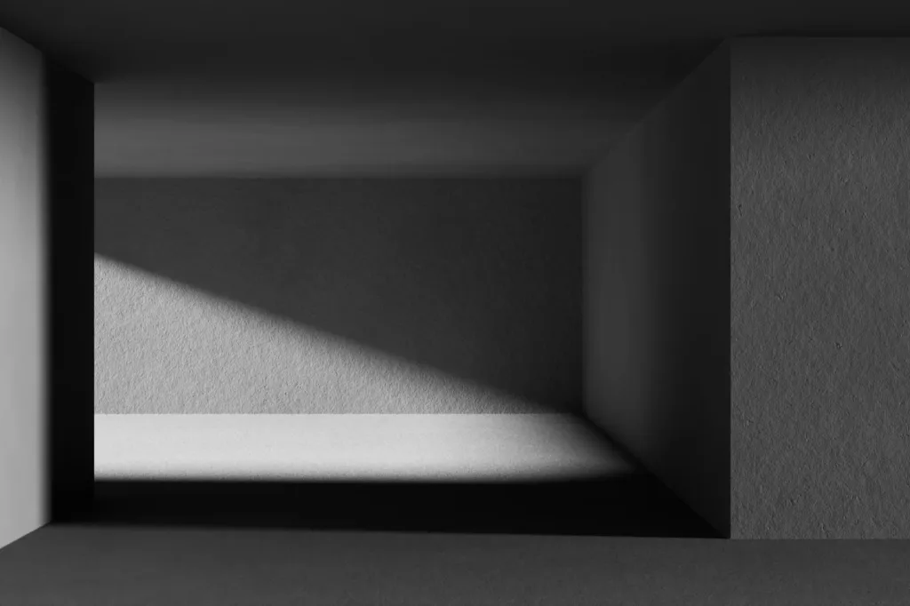 An empty concrete room in black and white, with light falling through the opening onto the back wall and floor