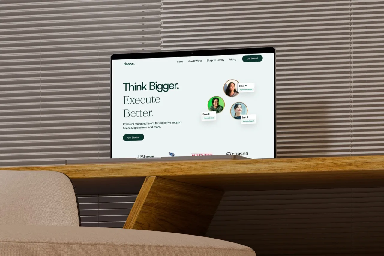 The Hire Donna homepage, Think Bigger and Execute Better, on a MacBook Pro on a wooden desk by window blinds
