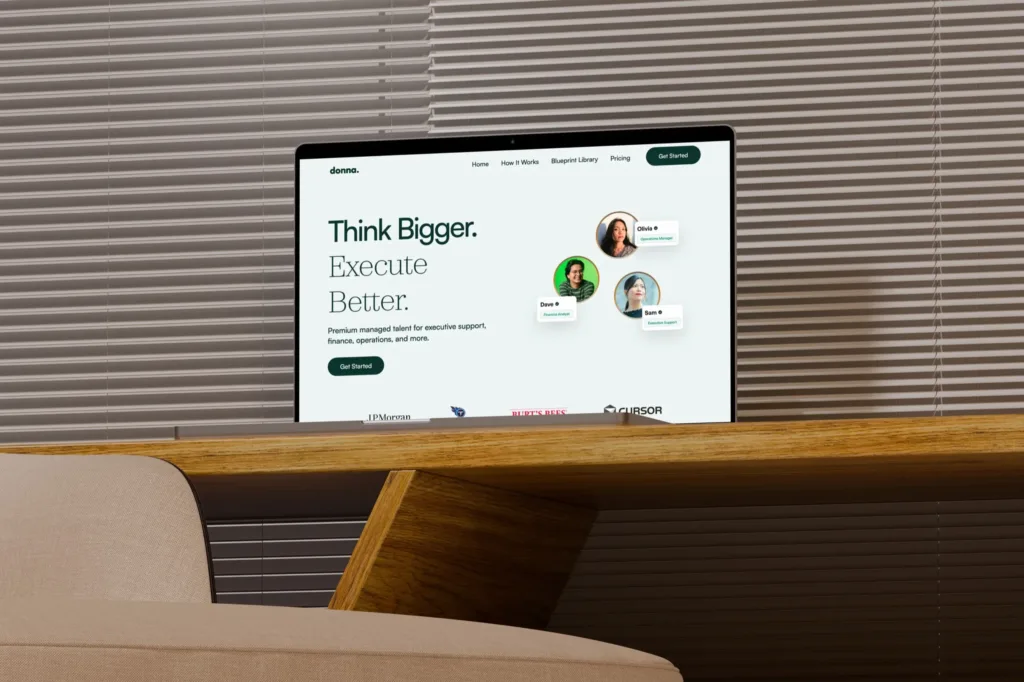 The Hire Donna homepage, Think Bigger and Execute Better, on a MacBook Pro on a wooden desk by window blinds