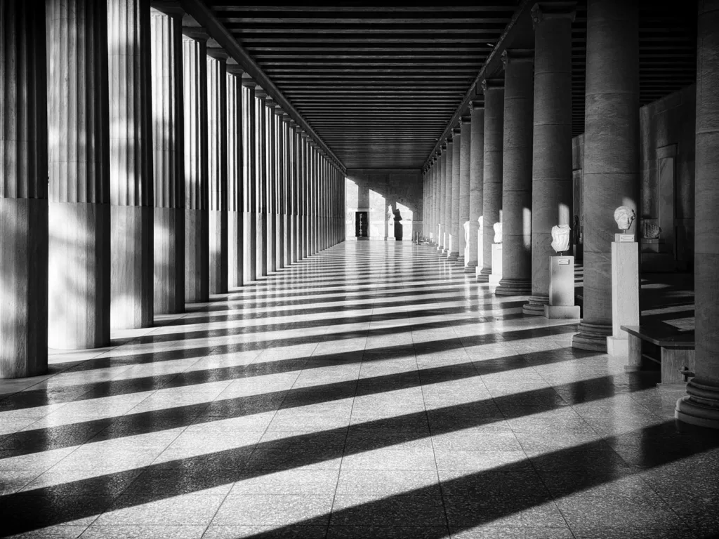 Sunlight falling in stripes between the columns of a long colonnade, in black and white