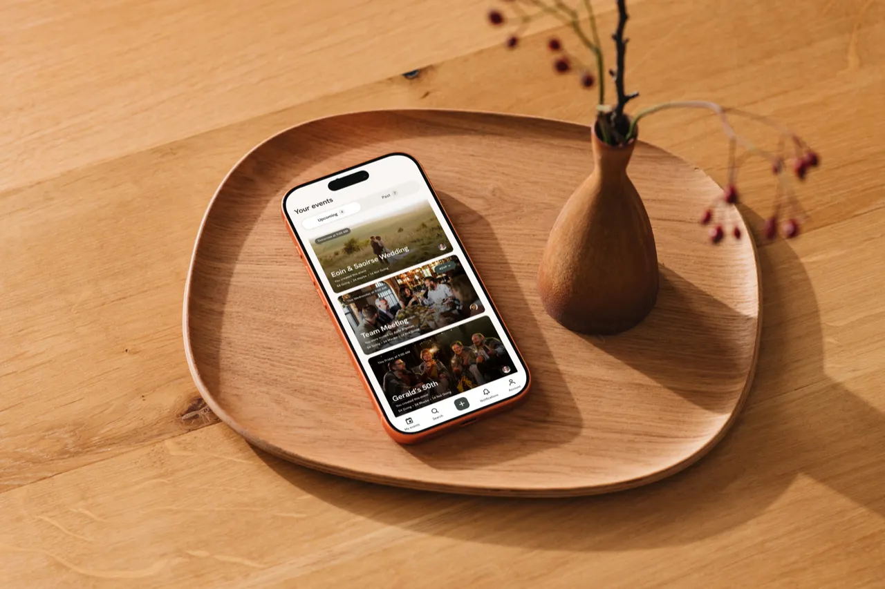The Cairn app's Your events screen, with a wedding, a team meeting and a 50th birthday, on an iPhone on a wooden tray beside a vase of rosehips