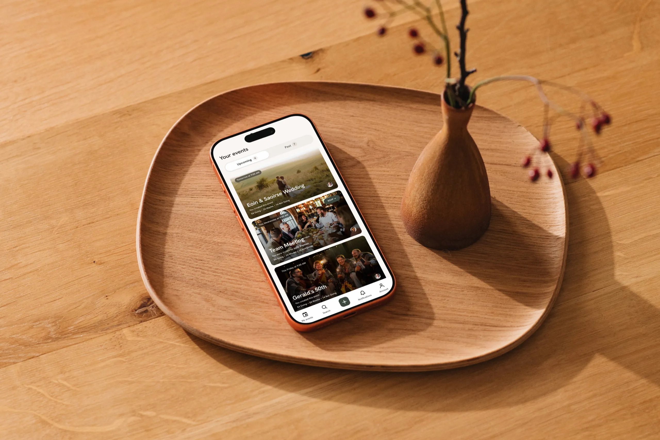 The Cairn app's Your events screen, with a wedding, a team meeting and a 50th birthday, on an iPhone on a wooden tray beside a vase of rosehips