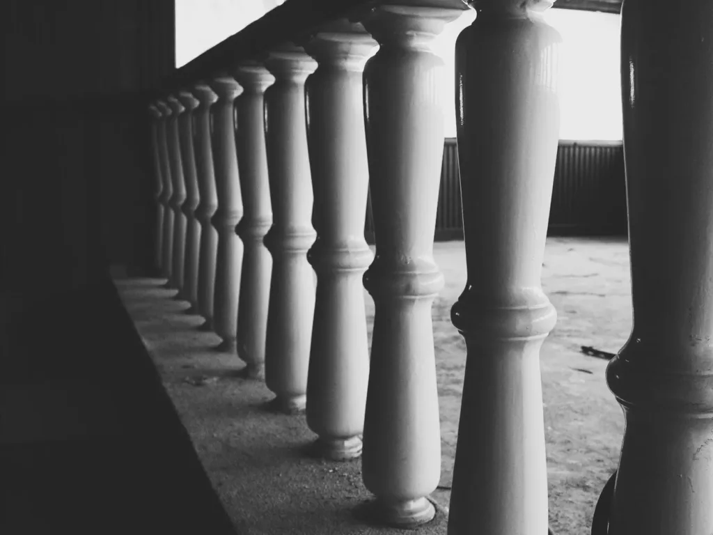 A row of white balusters receding into the distance, the nearest one cut off by the edge of the frame, in black and white