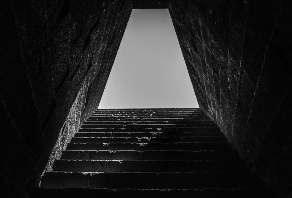 Stone steps rising through a dark passage towards a bright opening, in black and white